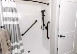 A white walk-in shower with grab bars on the walls, a handheld showerhead, and a striped shower curtain partially drawn to the side. A beige towel hangs on a towel bar outside the shower, and a white door with black hardware is visible to the right.