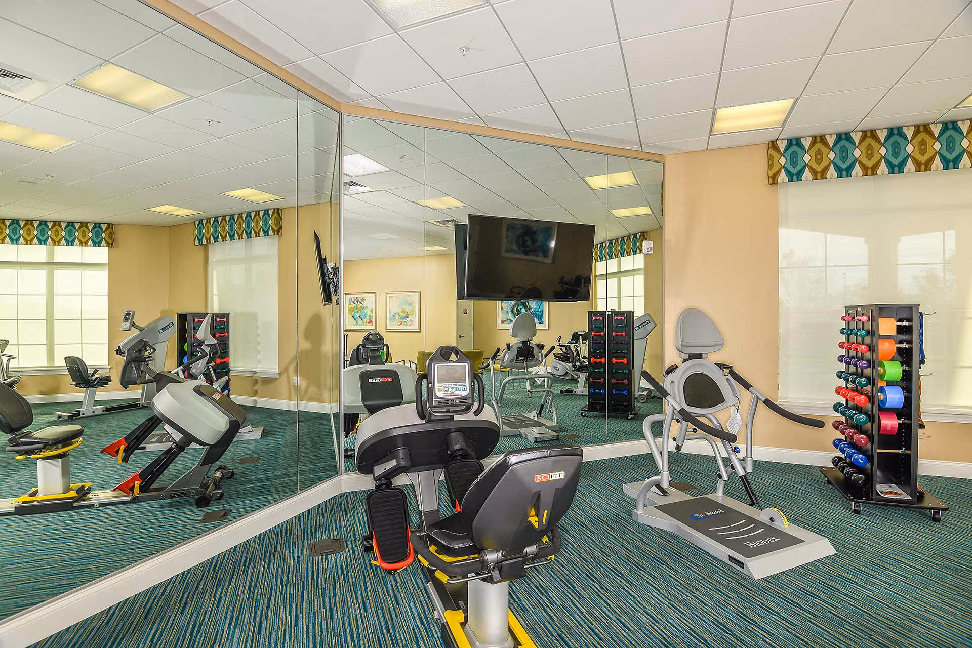 A fitness room with exercise equipment including stationary bikes, a treadmill, and a rack of colorful dumbbells. The room has large mirrors on one wall, windows with patterned valances, and a wall-mounted TV.