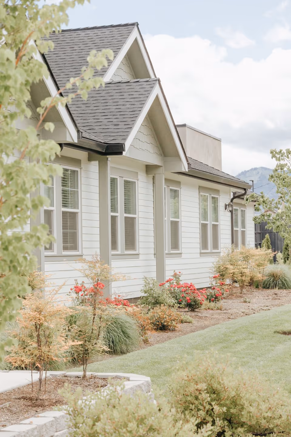 Exterior side view of a light-colored building with multiple windows, a dark shingled roof, and a landscaped garden featuring bushes, small trees, and red flowers under a partly cloudy sky.
