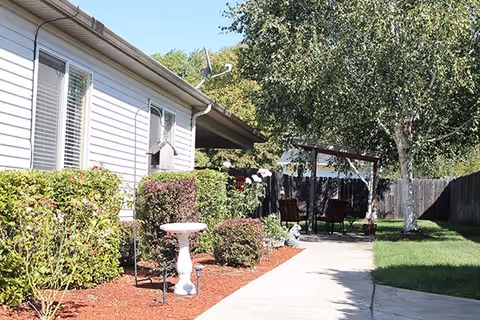 Outdoor garden area at the Douglas facility with a concrete pathway, trimmed bushes, a birdbath, a birdhouse mounted on the building, and a shaded seating area with chairs under a wooden pergola surrounded by trees and grass.