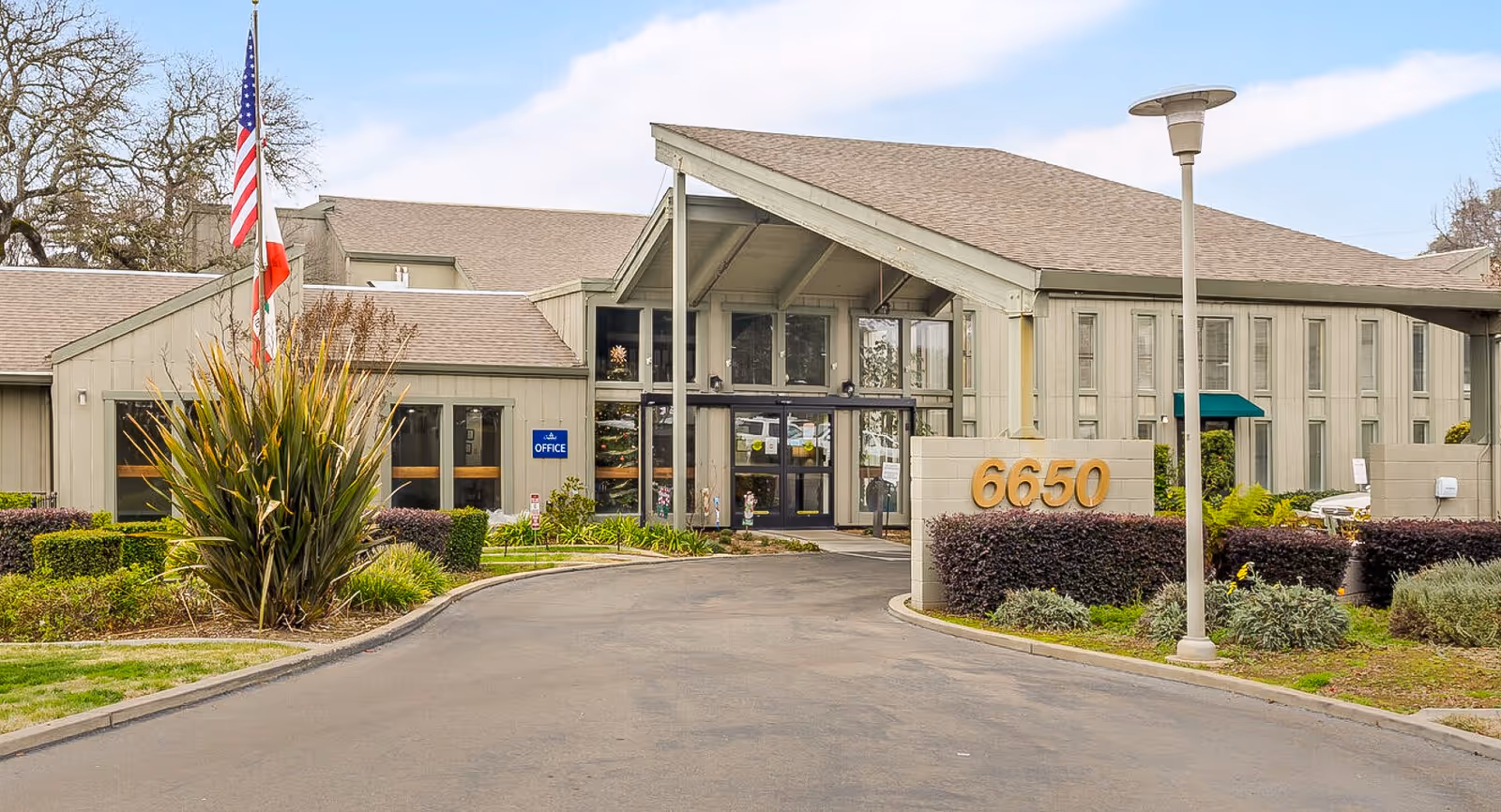 Exterior view of The Crest at Citrus Heights senior living facility showing the main entrance with a driveway, landscaped bushes, an American flag, and the building number 6650 displayed on a low wall.
