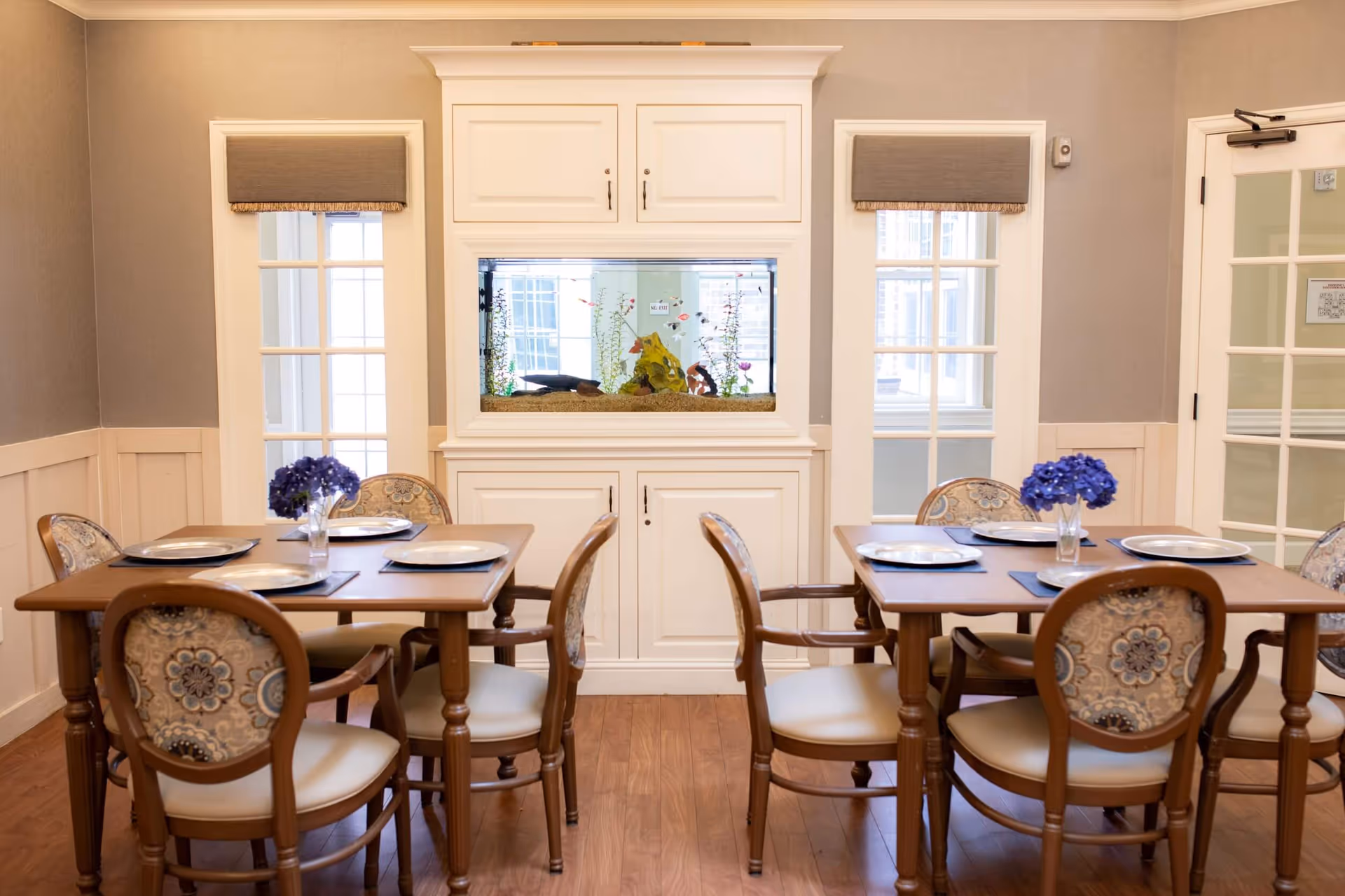 Bright dining room with two wooden tables and upholstered chairs set with plates, a built-in aquarium in white cabinetry, and small vases of purple flowers.