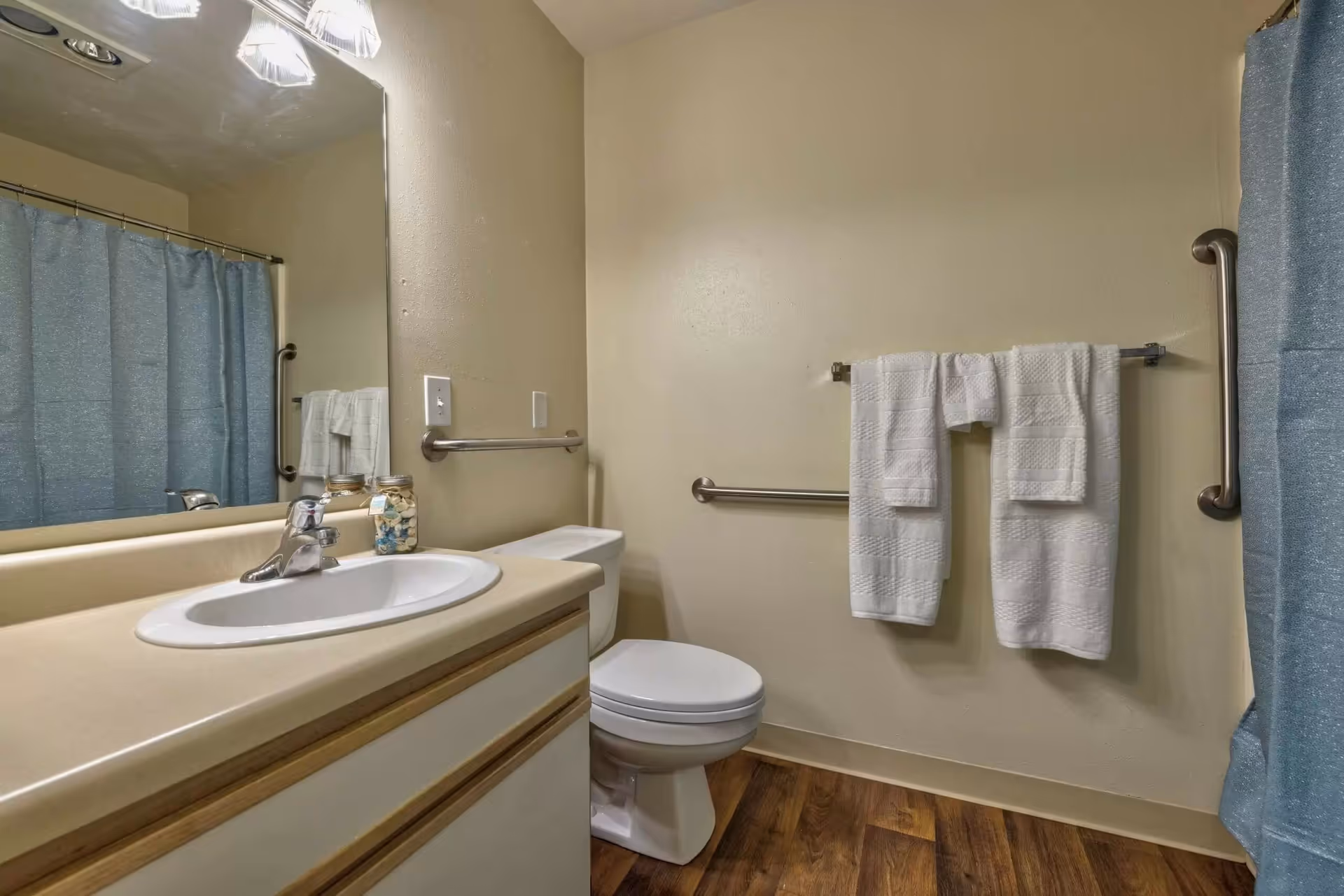 A clean bathroom with a sink and countertop on the left, a toilet in the center, and a shower with a blue curtain on the right. There are two white towels hanging on a towel rack on the wall, and grab bars are installed near the toilet and shower for accessibility. The floor has a wood-like finish.