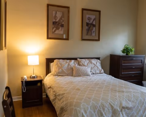 A neatly made bedroom with a bed, nightstand and lamp, framed artwork on the wall, and a dresser with a plant.