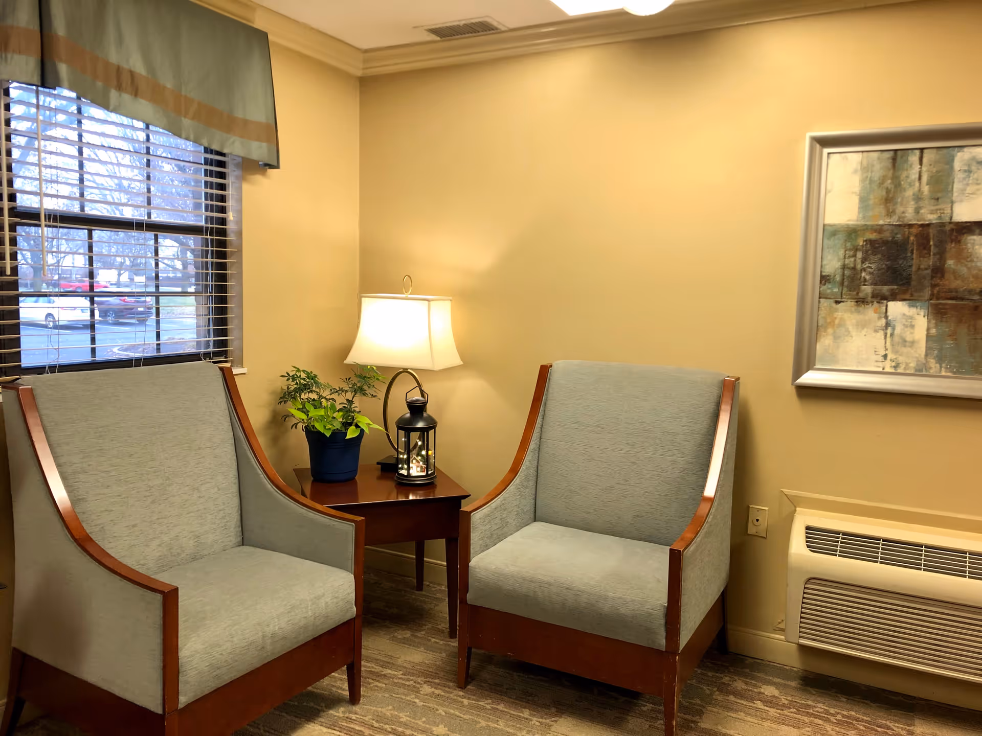A cozy seating area with two light gray upholstered armchairs with wooden frames, a small wooden side table between them holding a table lamp and a potted plant. There is a window with blinds and a green valance on the left side, a framed abstract painting on the beige wall, and a wall-mounted air conditioning unit below the painting.