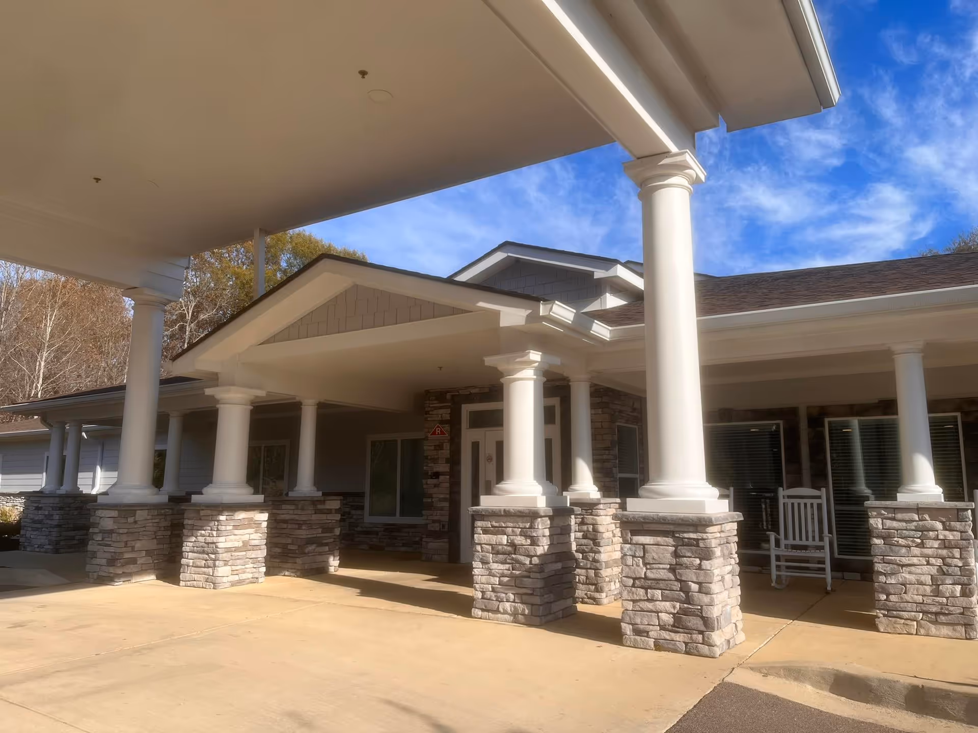 Exterior view of a senior living facility entrance with white columns on stone bases supporting a covered driveway. There is a white rocking chair near the entrance and trees in the background under a blue sky with some clouds.