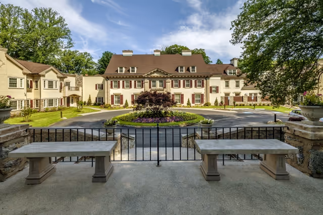 Front view of a large multi-story residential building with a circular driveway, landscaped center island, and stone benches in the foreground.