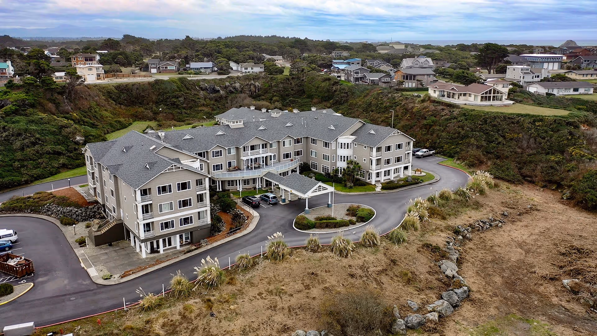 Aerial view of a large senior living community building surrounded by a circular driveway, with multiple cars parked near the entrance. The building is situated on a grassy and rocky terrain with other residential houses and greenery in the background under a cloudy sky.