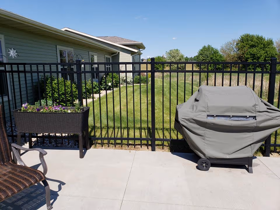 Outdoor patio area with a black metal fence, a covered grill, a planter box with flowers, and a brown wicker chair. Behind the fence is a grassy area and a building with green siding and windows. Trees and a clear blue sky are visible in the background.