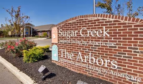 Brick entrance sign reading 'Sugar Creek' and 'The Arbors' in front of a senior living building with landscaping under a clear sky.