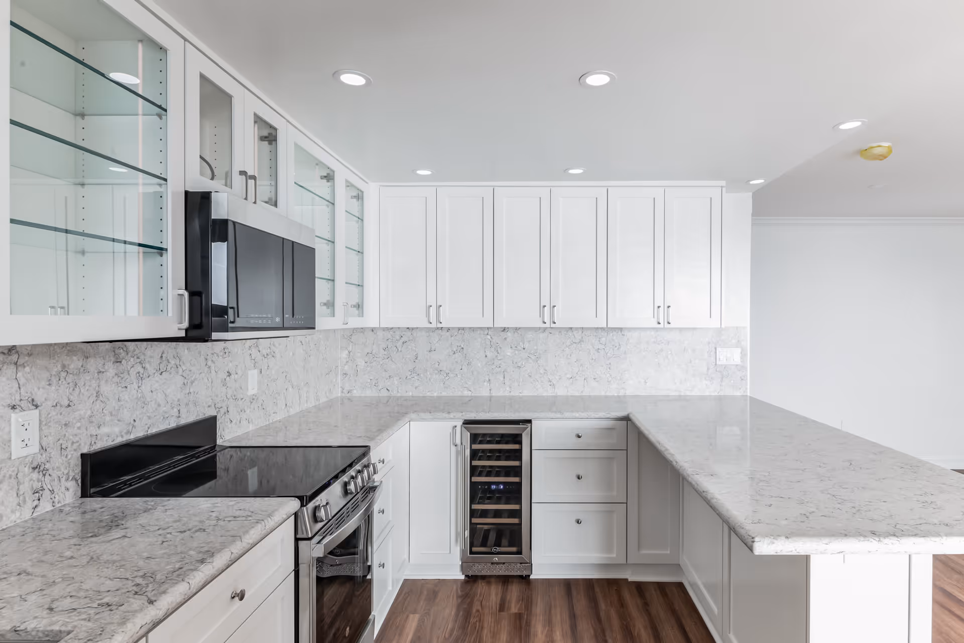 Modern kitchen with white cabinetry, marble countertops, glass-front upper cabinets, a built-in microwave, an electric stove, and a wine cooler. The floor is wood, and recessed lighting is installed in the ceiling.