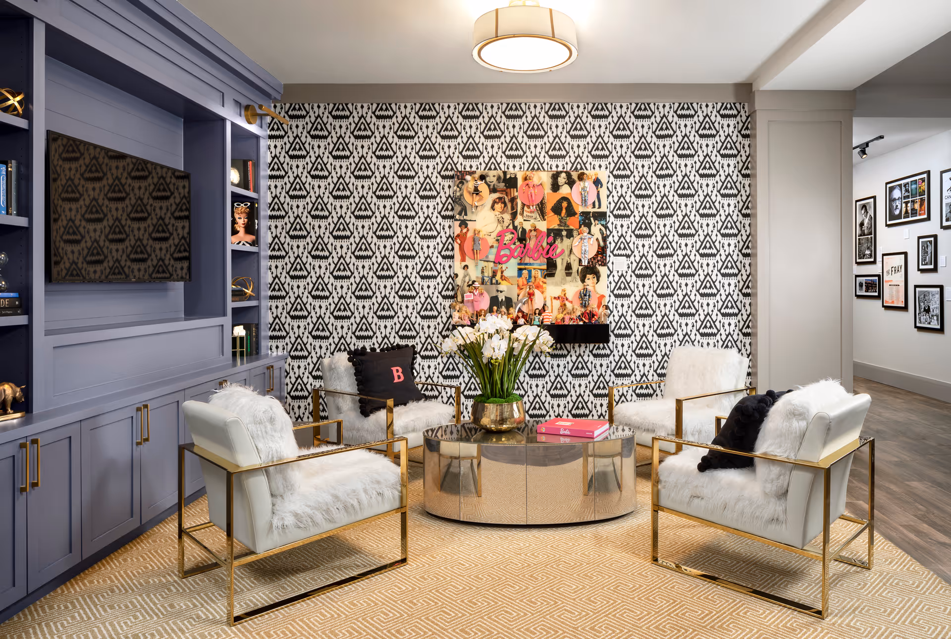 Stylish lounge with four white fur-upholstered chairs around a round coffee table, patterned wallpaper, and built-in shelving with a TV.