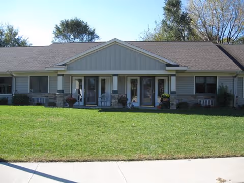 Single-story building with a gray roof and light gray siding, featuring two front doors with small porches and chairs, surrounded by a green lawn and trees in the background under a clear sky.