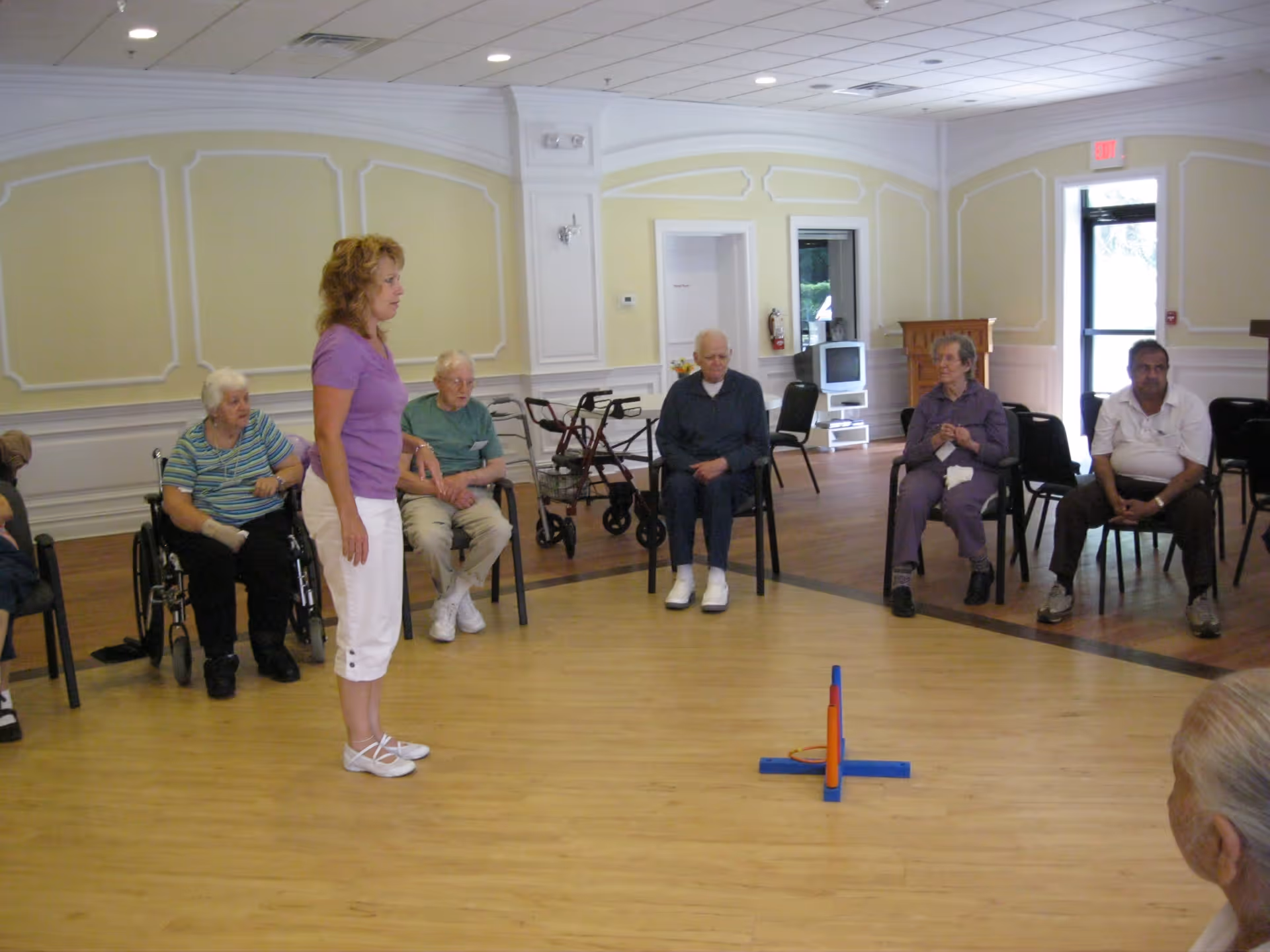 A group of elderly people seated in a semi-circle in a spacious room with wooden floors and pale yellow walls with white trim. A woman in a purple shirt and white pants stands in the center, possibly leading an activity. Some of the elderly individuals are seated in chairs, one in a wheelchair, and there are walkers visible in the background. A small game or activity setup is on the floor in the middle of the room.
