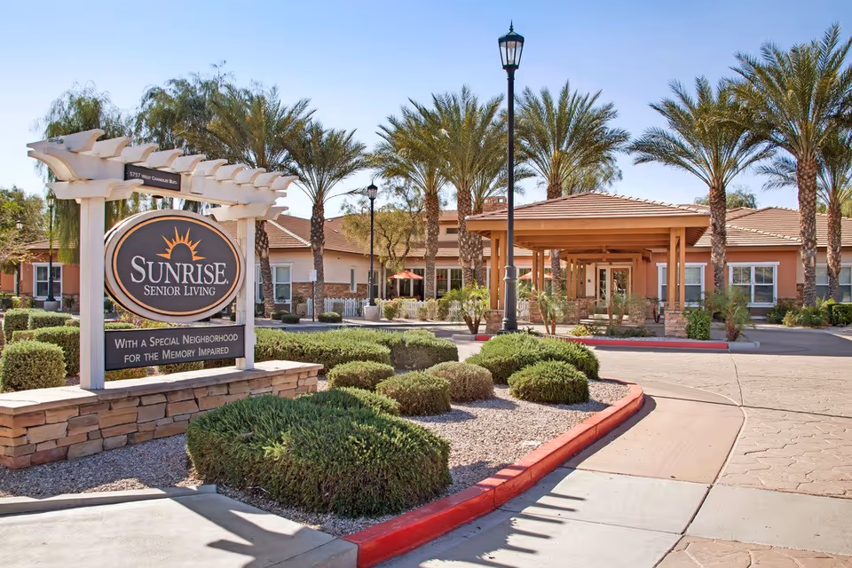 Front entrance of the Sunrise Senior Living facility with a large sign, palm trees, shrubs, and a covered porte-cochère.