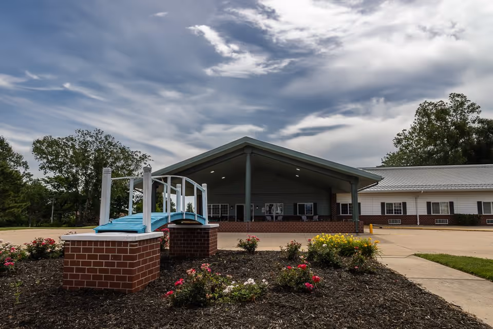 Exterior view of Bailey Pointe at Plattsmouth facility showing a covered entrance with a peaked roof, a small decorative blue bridge over a flower bed with blooming flowers, and a paved driveway. Trees and a partly cloudy sky are visible in the background.