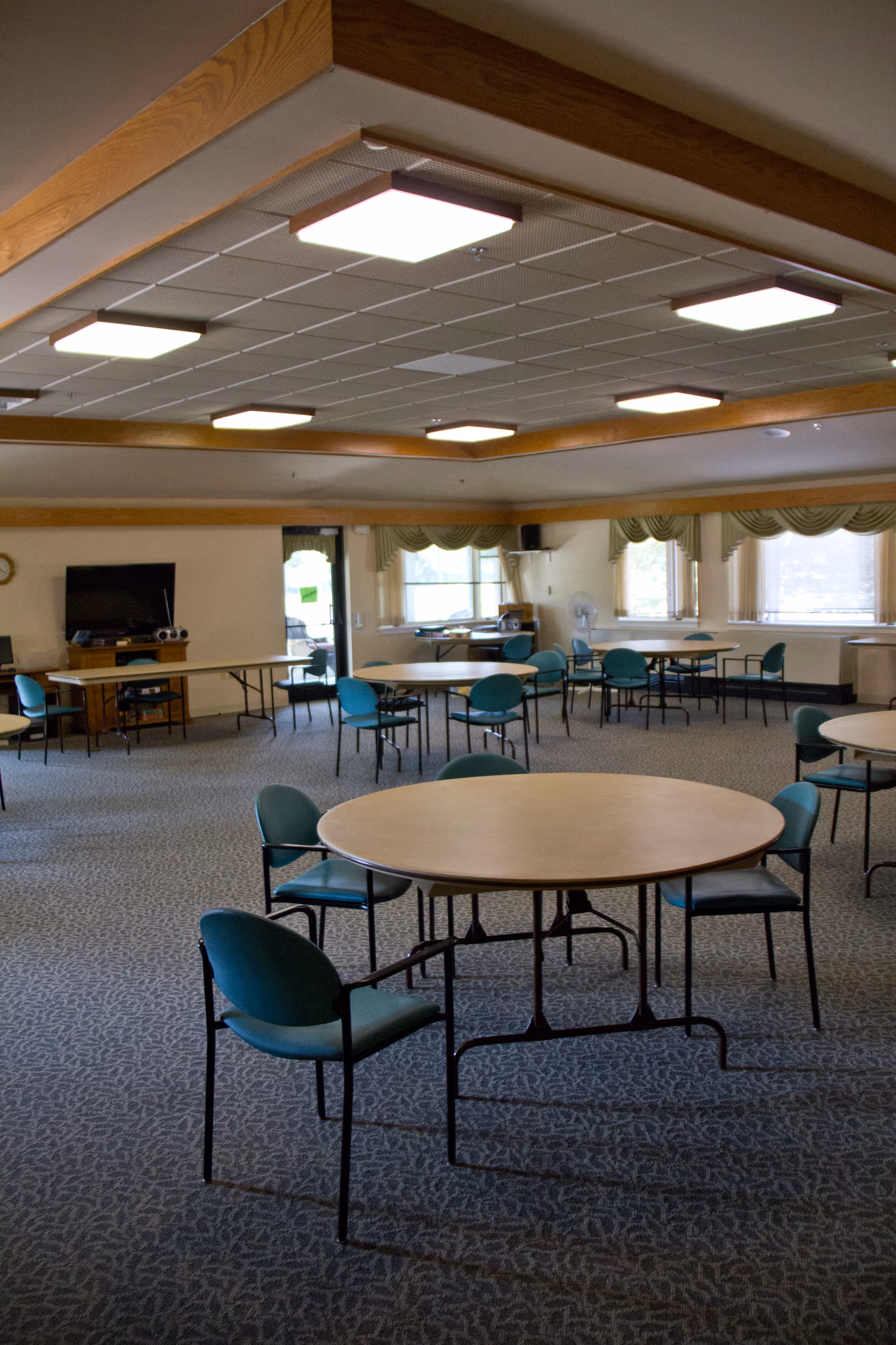 A spacious room with multiple round tables and teal chairs arranged around them. The room has a carpeted floor, large windows with valance curtains, and a ceiling with recessed lighting. There is a television and some equipment on a table against one wall.