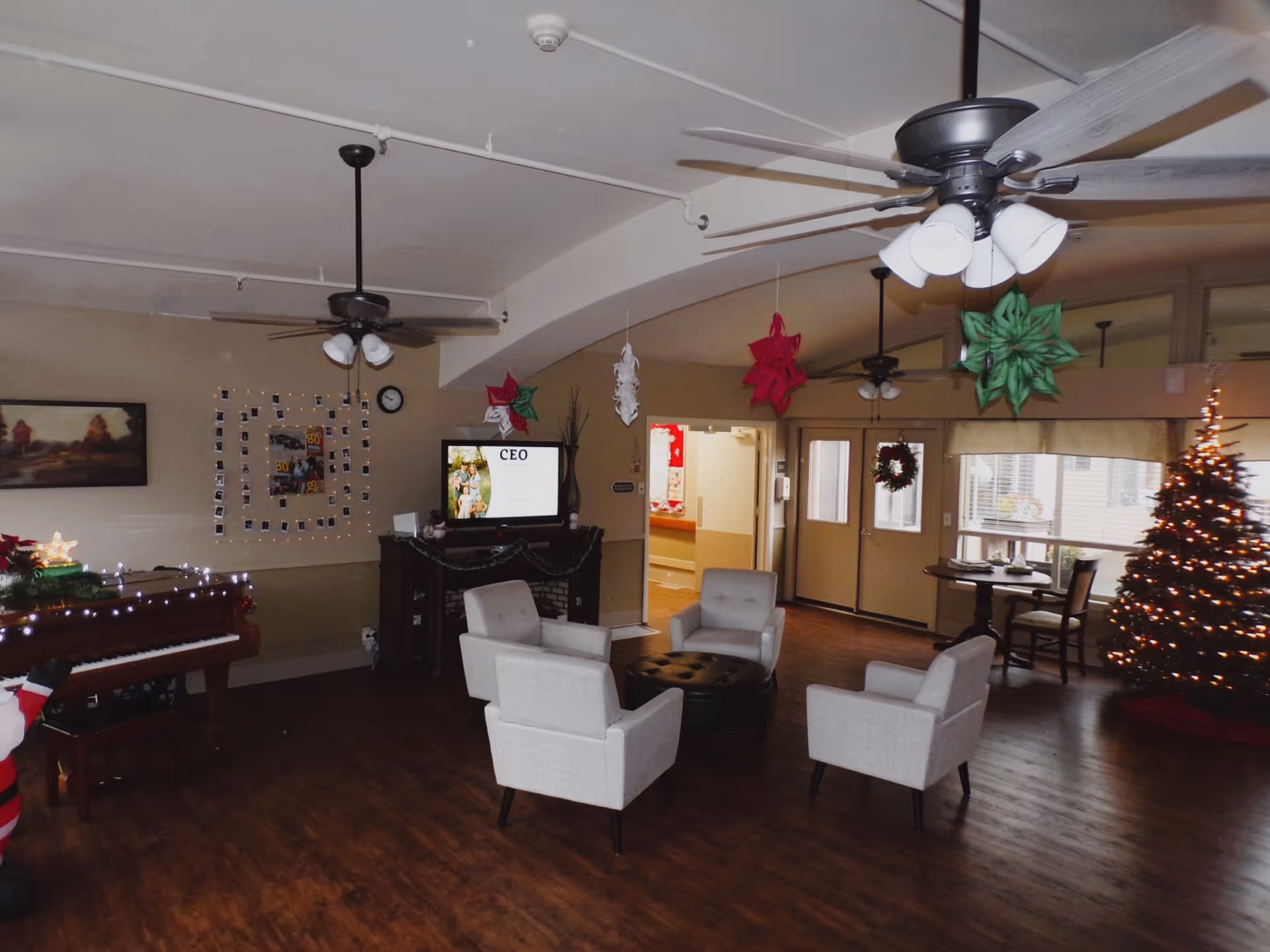 A cozy living room area in a senior living facility decorated for the holidays with a Christmas tree, hanging paper star decorations, and a piano with string lights. The room has four light gray armchairs arranged around a round black ottoman, ceiling fans with lights, a TV on a stand, and a small dining table near windows.