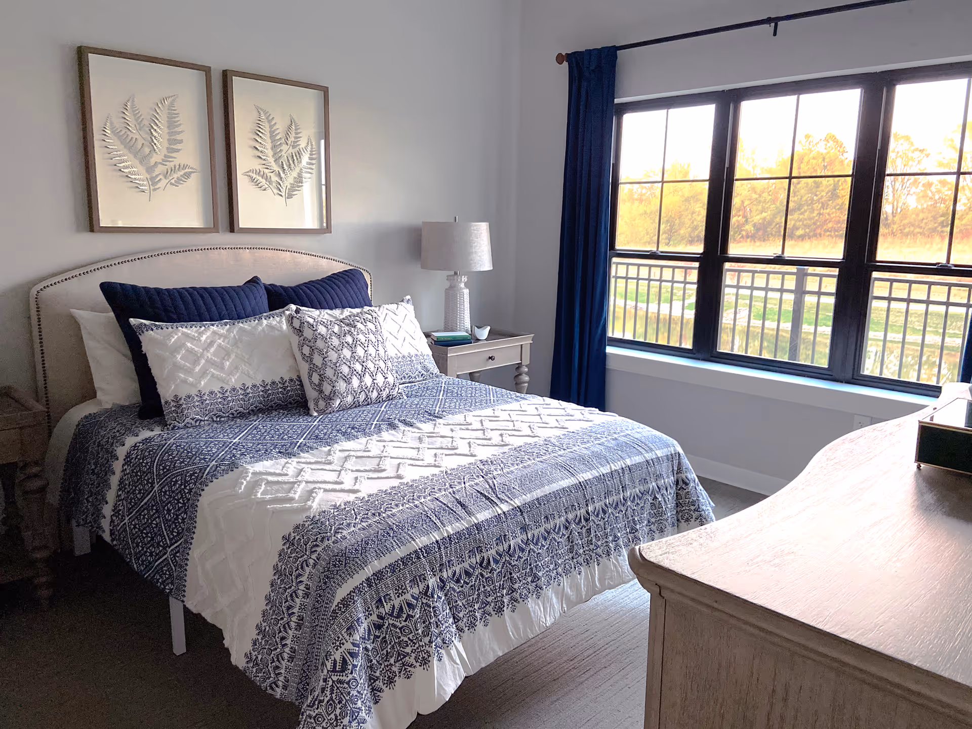 Sunlit bedroom with a blue-and-white patterned bed, bedside table and lamp, large window with dark curtains, and a wooden dresser.
