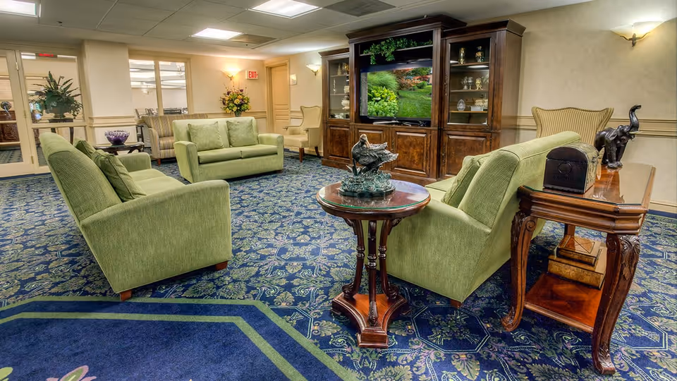 A cozy senior living facility lounge area with green upholstered sofas and armchairs arranged around a wooden coffee table and a large wooden entertainment center with a TV displaying a garden scene. The room features patterned blue carpet, beige walls, decorative plants, and warm lighting.