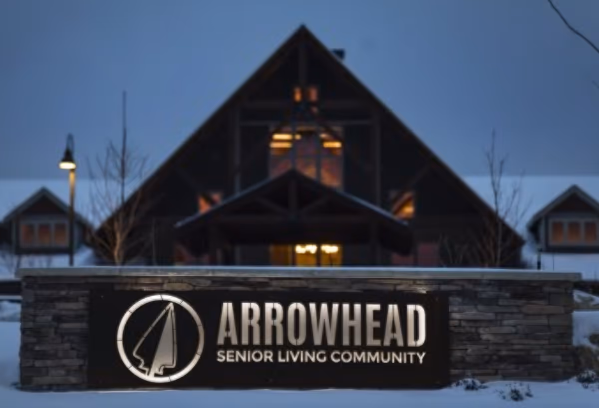 Illuminated sign for Arrowhead Senior Living Community in front of a large building with a peaked roof at dusk or night, with snow on the ground.