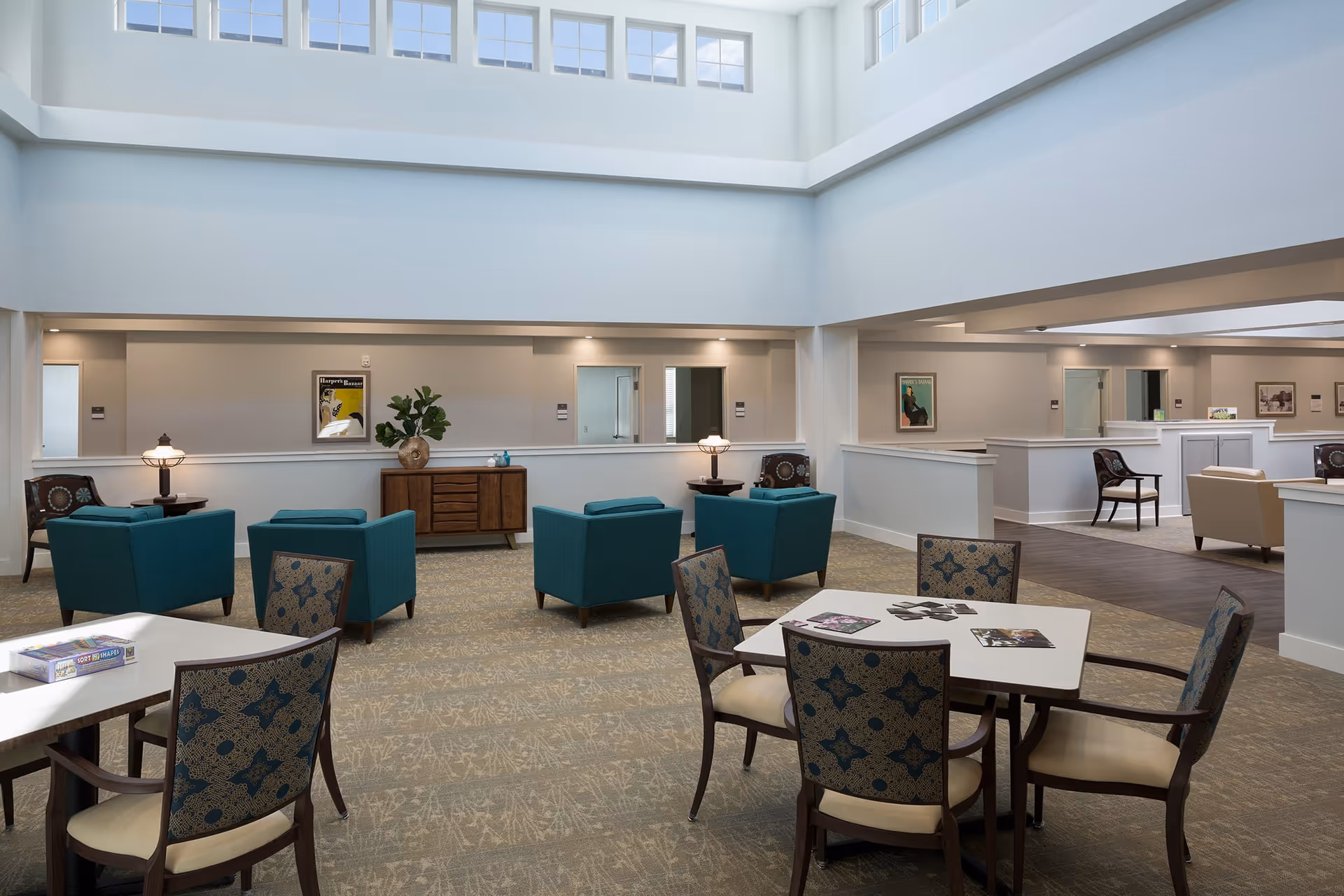 A spacious and well-lit common area in a senior living facility with high ceilings and large windows. The room features several teal armchairs arranged in small groups, patterned chairs around square tables, a wooden sideboard with a plant and decorative items, and framed artwork on the walls. The carpeted floor and soft lighting create a comfortable and inviting atmosphere.