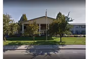 Front view of a two-story senior living building with a covered entrance, flagpole, manicured lawn, and trees.