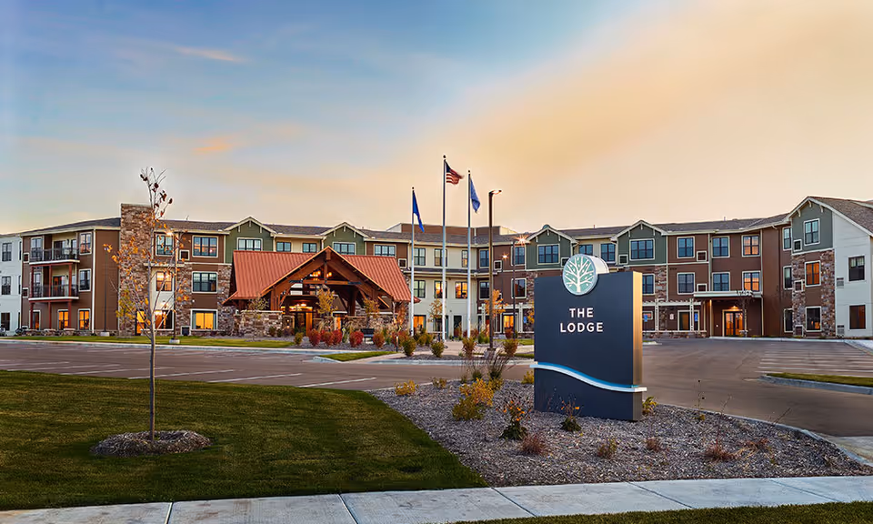 Exterior view of a senior living facility named The Lodge at sunset, featuring a three-story building with multiple windows, a covered entrance with wooden beams, three flagpoles with flags, a parking lot, and landscaped grounds with grass and small plants.