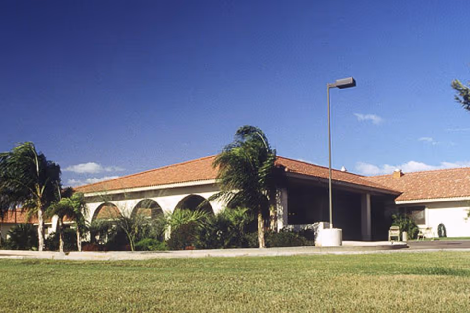 Exterior view of Desert Cove Nursing Center showing a single-story building with a red-tiled roof, arched windows, palm trees, a lamp post, and a well-maintained grassy lawn under a clear blue sky.