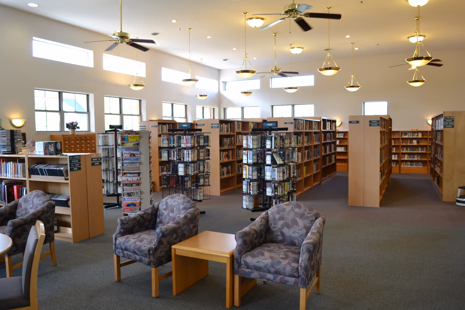 Interior view of a library room with multiple bookshelves filled with books and media. There are two patterned armchairs and a small wooden table in the foreground. The room is well-lit with large windows, ceiling fans, and hanging light fixtures.