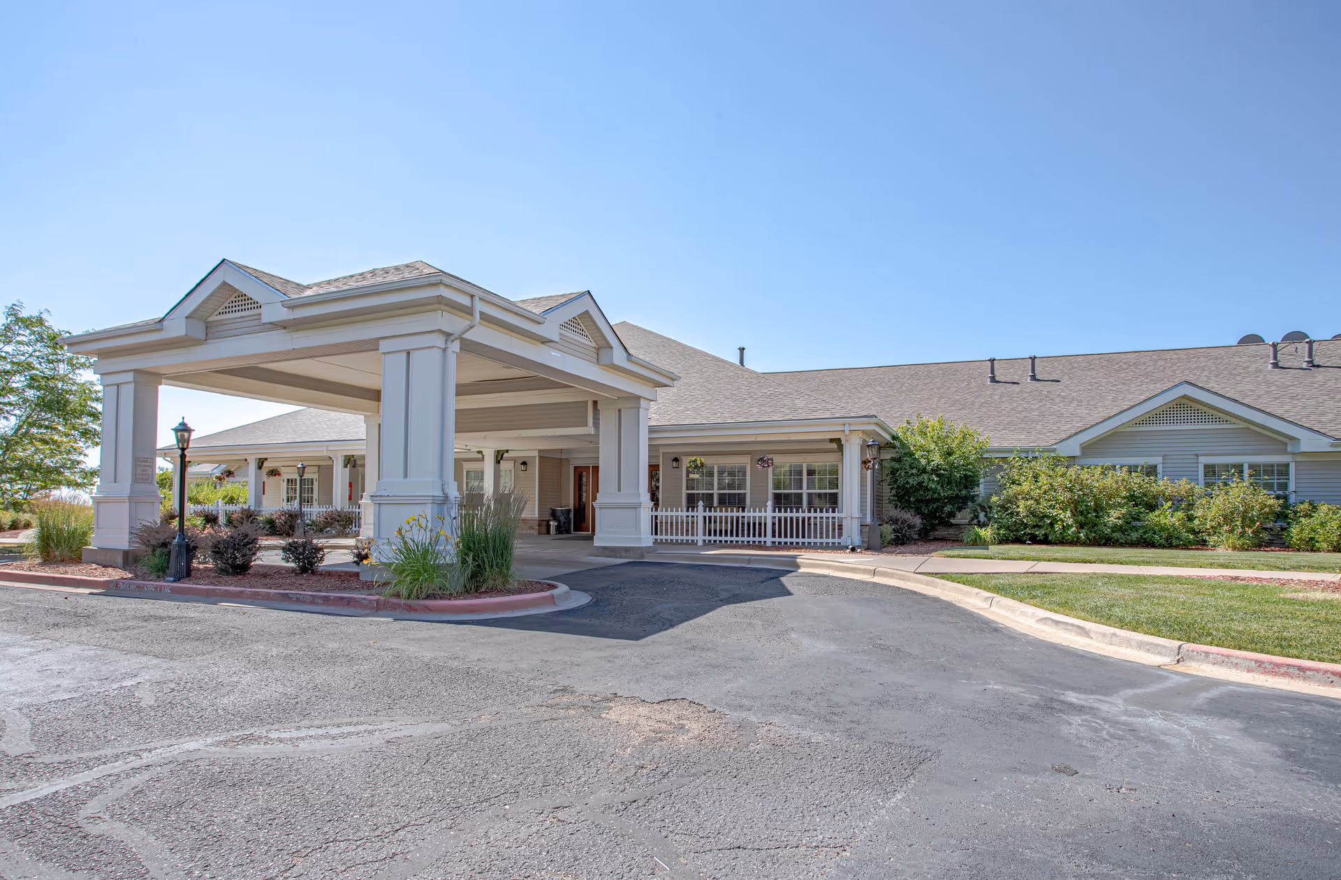 Front exterior view of Bear Creek Assisted & Senior Living facility showing a covered entrance with white pillars, a driveway, landscaped bushes, and a clear blue sky.