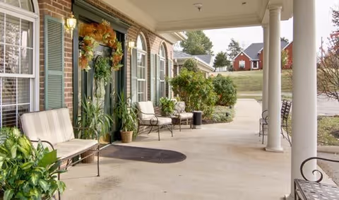 Covered outdoor patio area at Dogwood Pointe Senior Living with benches and chairs along the wall, potted plants, and a green door decorated with a fall wreath. The area is supported by white columns and overlooks a grassy area with trees and a red building in the background.