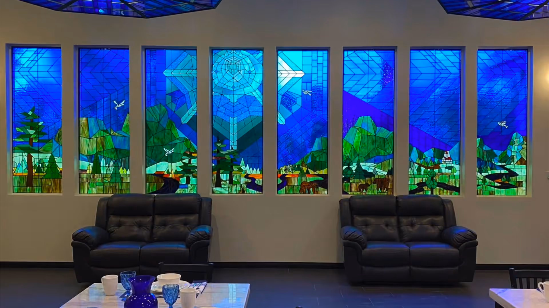 Interior view of a common area with two black leather sofas facing a table with cups and glasses. Behind the sofas are eight tall stained glass windows depicting a colorful nature scene with trees, mountains, and birds under a blue sky.