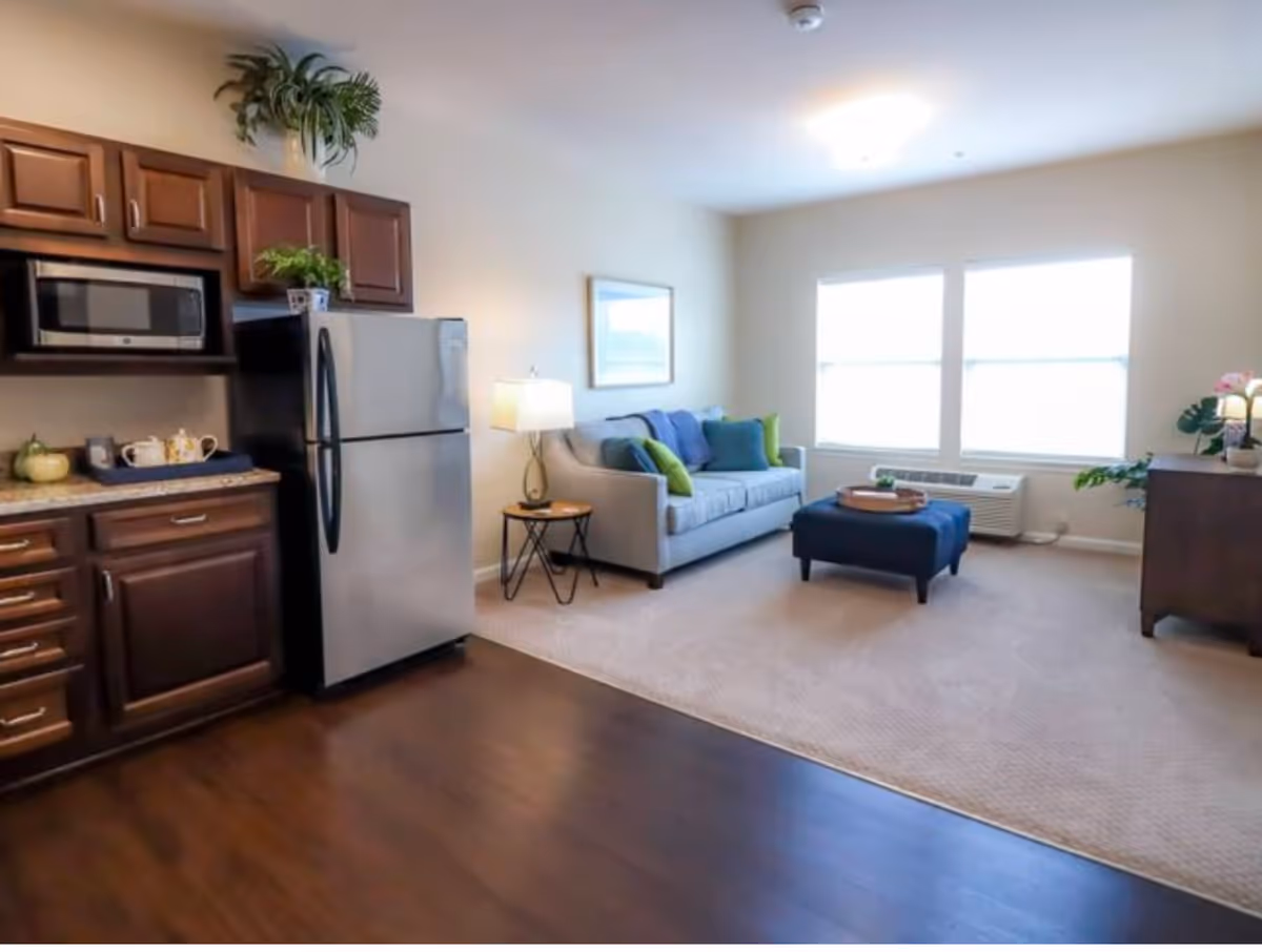 Open-plan apartment interior showing a kitchenette with a stainless refrigerator and a sitting area with a sofa, ottoman, and large windows.