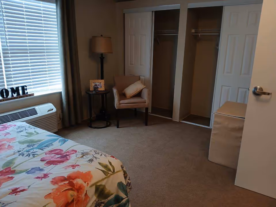 A bedroom with a floral bedspread partially visible in the foreground, a window with blinds and curtains, a small round side table with a framed photo and a lamp, a beige armchair with a cushion, and two open closets with sliding doors. There is also a beige storage ottoman near the door.