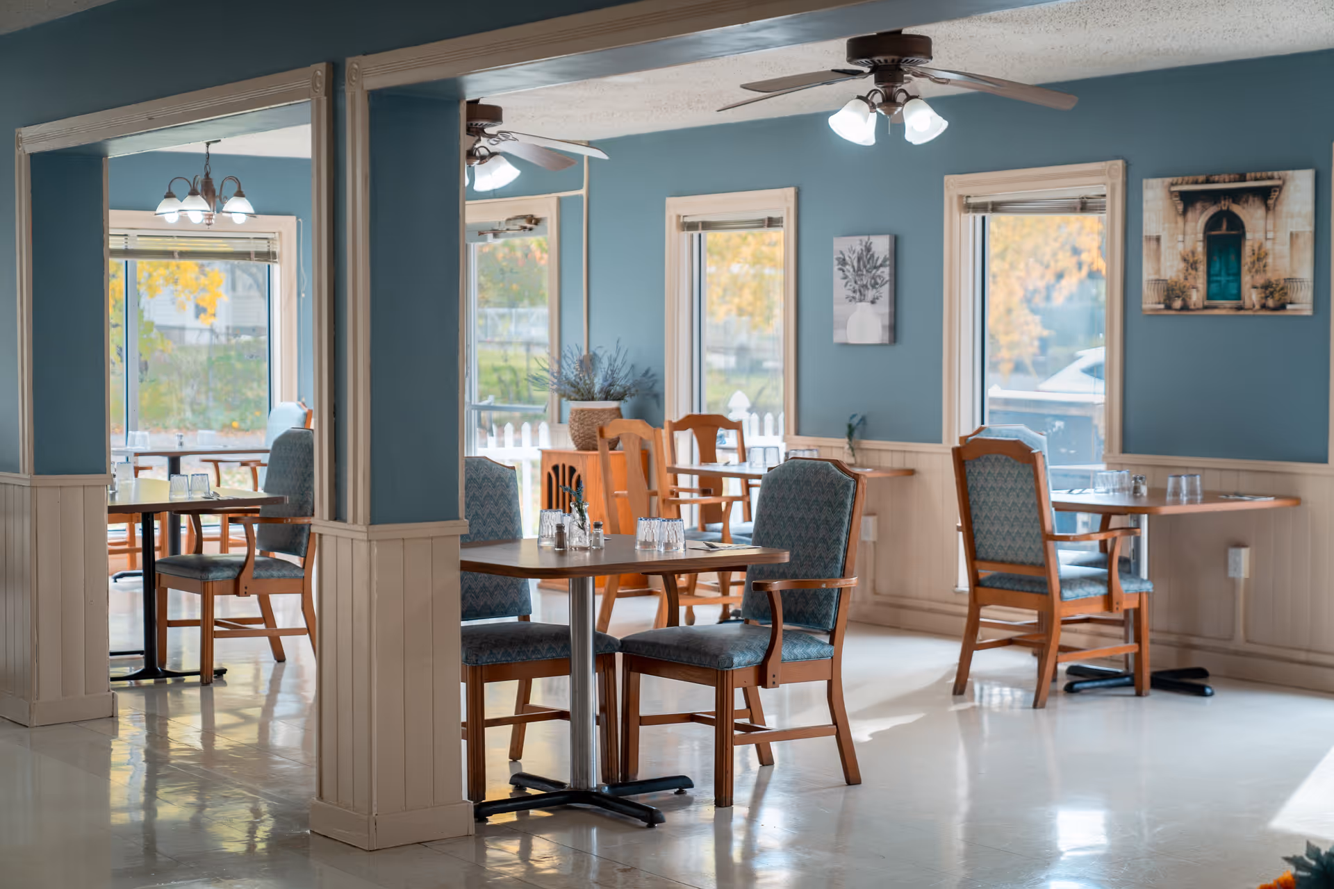Sunlit dining room with wooden tables and upholstered chairs, ceiling fans, and blue walls.