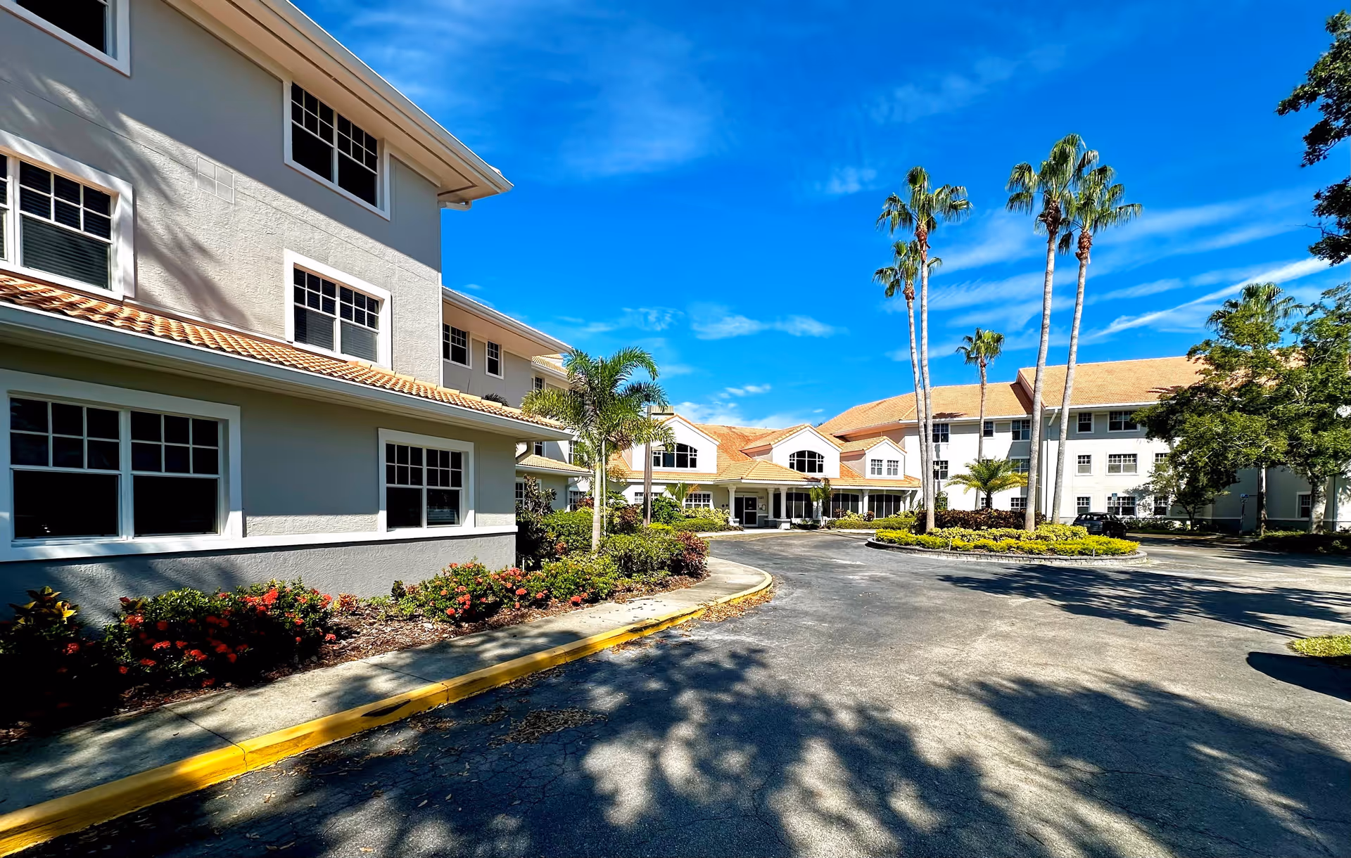 Exterior view of a senior living facility with a curved driveway, landscaped garden beds with flowers and palm trees, and a multi-story building with white walls and a tiled roof under a blue sky.