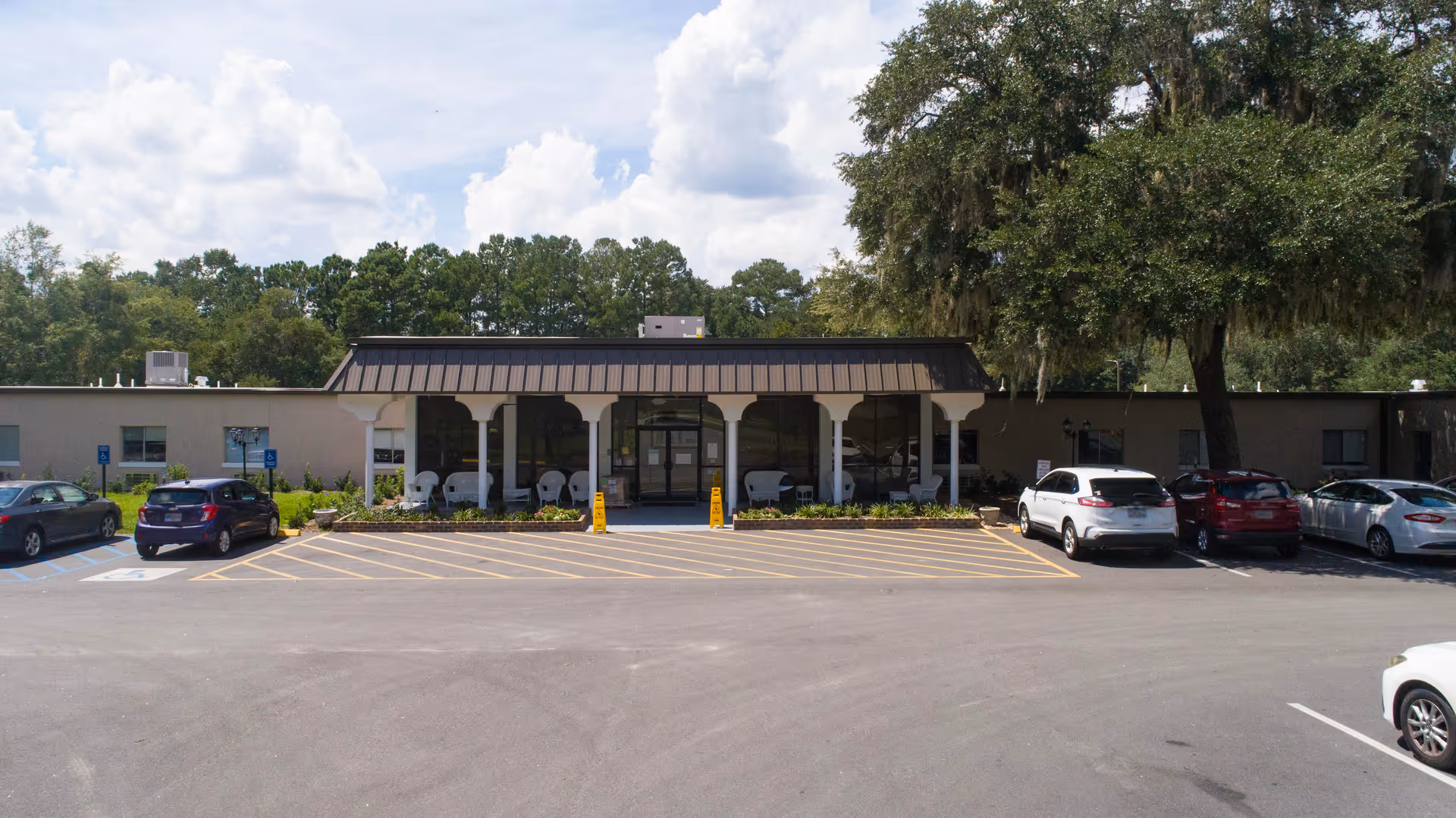 Front exterior view of Magnolia Manor of Midway, showing a single-story building with a covered entrance supported by white columns. Several cars are parked in the parking lot in front of the building, and there are trees and greenery surrounding the area under a partly cloudy sky.