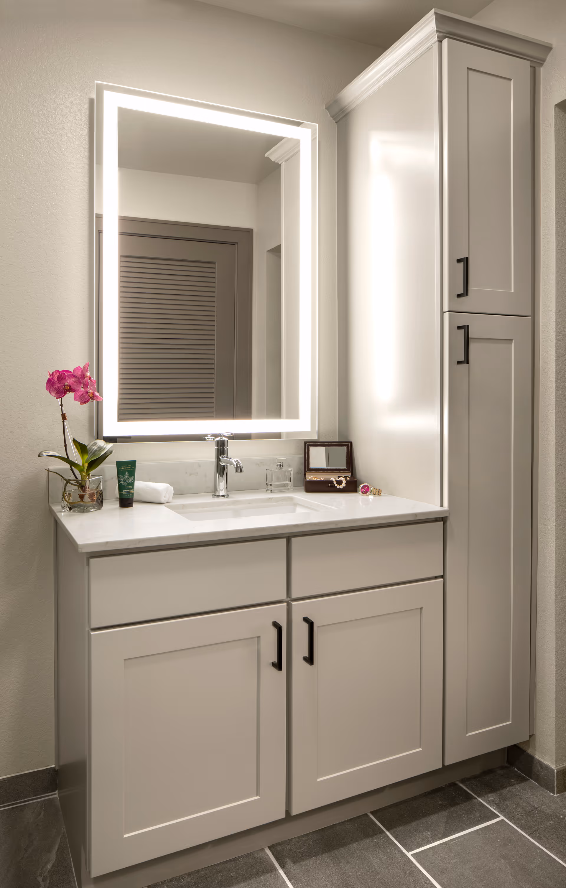 Modern bathroom vanity with an illuminated rectangular mirror, sink, countertop accessories, lower cabinets and a tall storage cabinet.