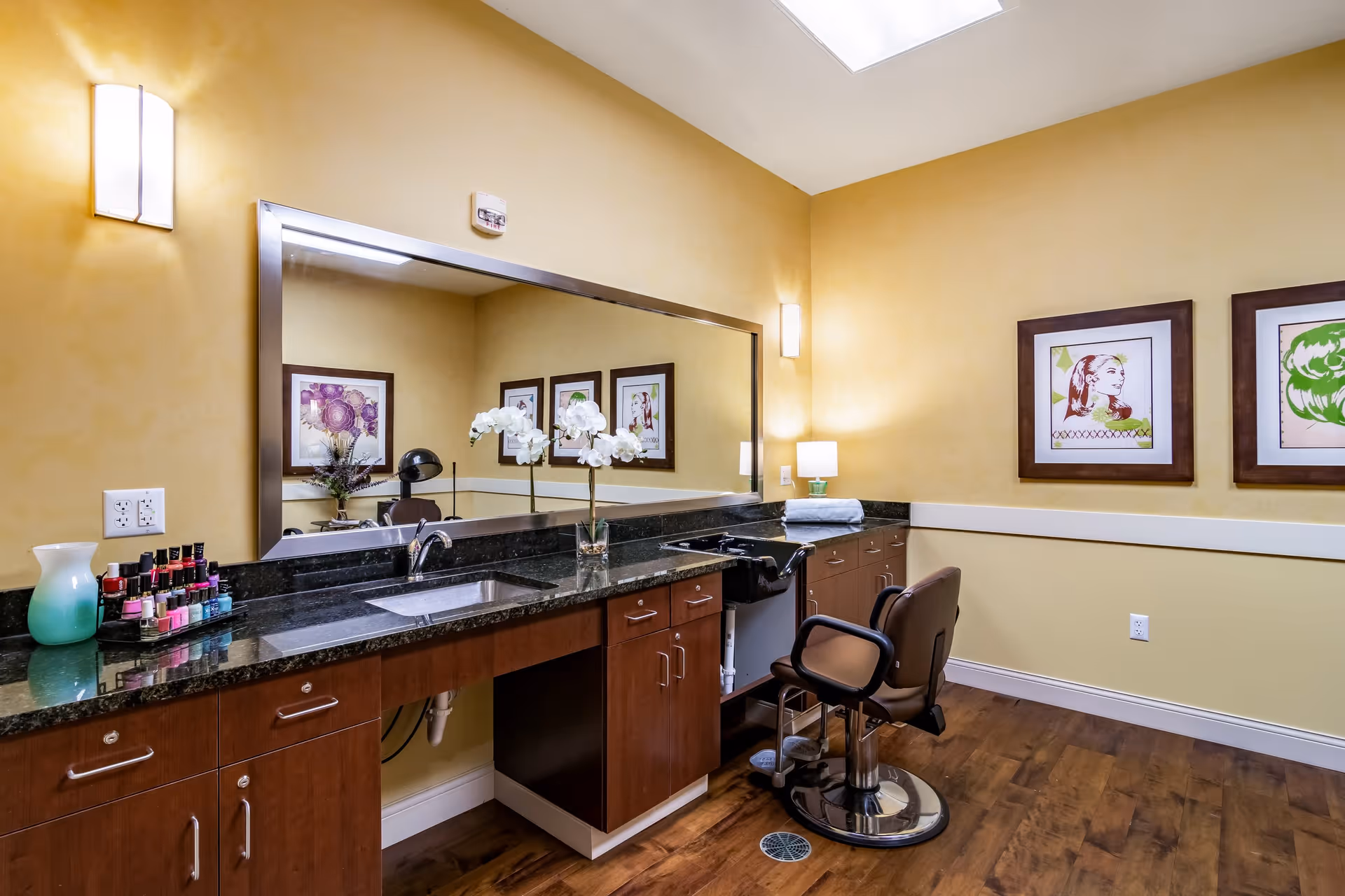 Interior view of a salon or grooming area with a large mirror above a black granite countertop, a salon chair, nail polish bottles on the counter, a vase with white flowers, and framed artwork on the yellow walls.