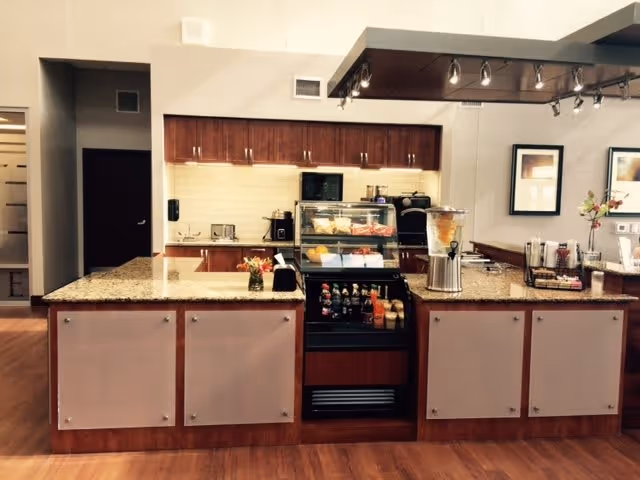 A modern kitchen area with granite countertops and wooden cabinets. The counter has a display case with various food items, a beverage dispenser, and a selection of bottled drinks. The background shows a tiled backsplash, a microwave, and other kitchen appliances. The floor is wooden, and there are framed pictures on the wall.