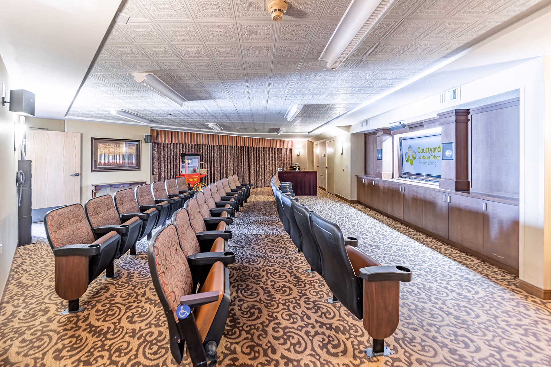 A senior living facility theater room with two rows of cushioned seats facing a large screen displaying the Courtyard at Mount Tabor Senior Living logo. The room has patterned carpet, a popcorn machine in the back corner, and decorative ceiling tiles with fluorescent lighting.