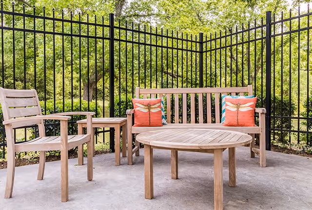 Outdoor seating area with wooden bench, two wooden chairs, a round wooden table, and a small wooden side table. The bench has two decorative pillows with dragonfly designs. The area is enclosed by a black metal fence and surrounded by green trees and bushes.