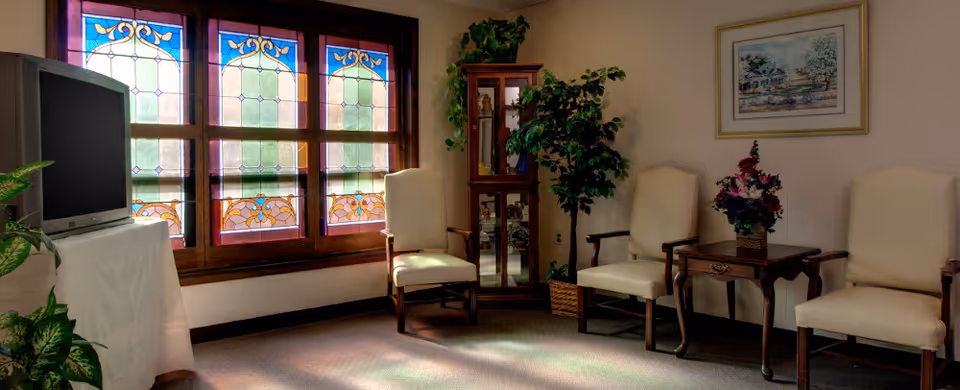 A cozy room with three cream-colored armchairs arranged around a wooden side table holding a floral arrangement. There is a stained glass window with colorful patterns letting in light, a TV on a stand covered with a white cloth, a tall wooden cabinet with glass doors, and a potted plant in the corner. A framed painting hangs on the wall.
