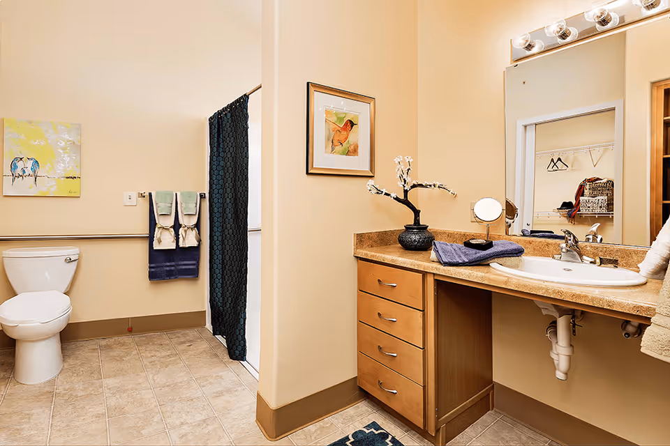 A clean and well-lit bathroom featuring a white toilet, a shower with a dark blue curtain, and a vanity with a sink, countertop, and wooden drawers. The countertop has a decorative vase with white flowers, a small round mirror, and folded towels. A large mirror is mounted above the sink, reflecting a closet with hangers and shelves. The walls are painted beige, and there is a painting of birds on the wall near the toilet.