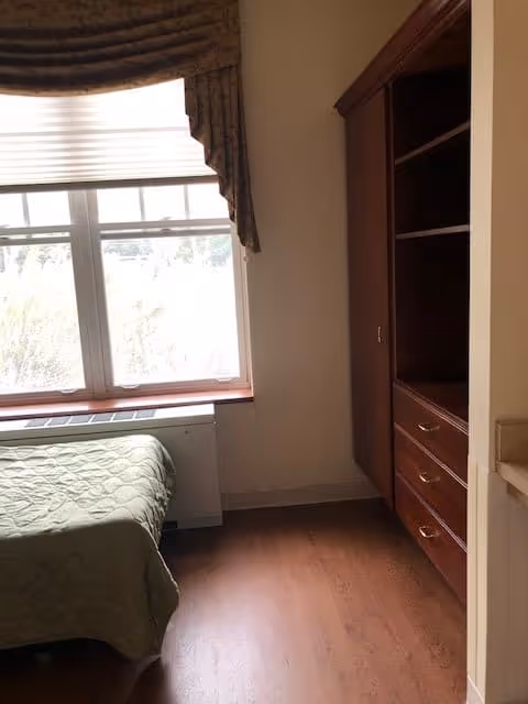A small bedroom with a single bed covered with a green quilt near a window with blinds and a valance. Next to the bed is a wooden built-in cabinet with shelves and drawers. The floor is wooden.
