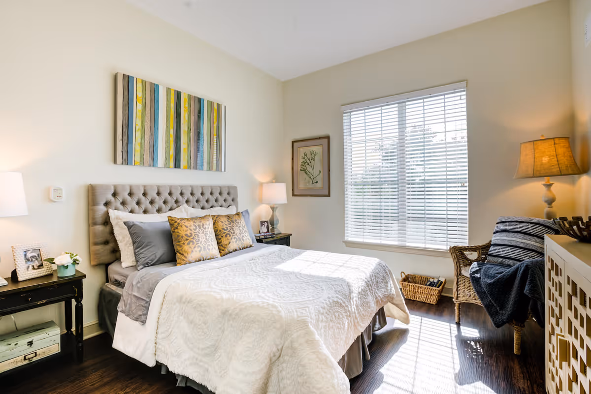 Sunlit bedroom with a tufted headboard, made bed with decorative pillows, bedside tables, a window with blinds, and a wicker chair.