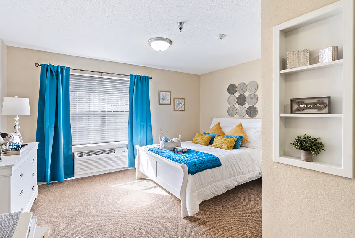 A bright and cozy bedroom with a white bed adorned with blue and yellow pillows and a blue throw blanket. The room features a large window with blue curtains, a white dresser with a lamp, and built-in wall shelves holding decorative items including a small plant and a framed sign. The walls are painted beige and the floor is carpeted.