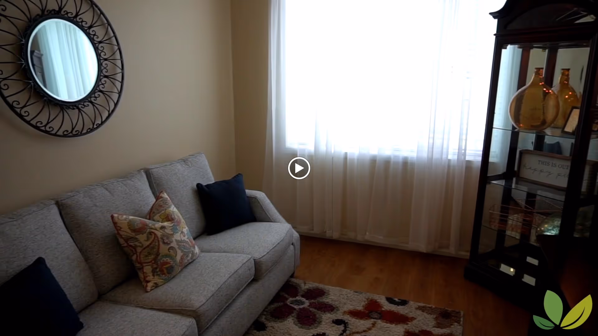 Cozy living room with a gray sofa, decorative pillows, round wall mirror, sheer curtains over a bright window, and a glass display cabinet.