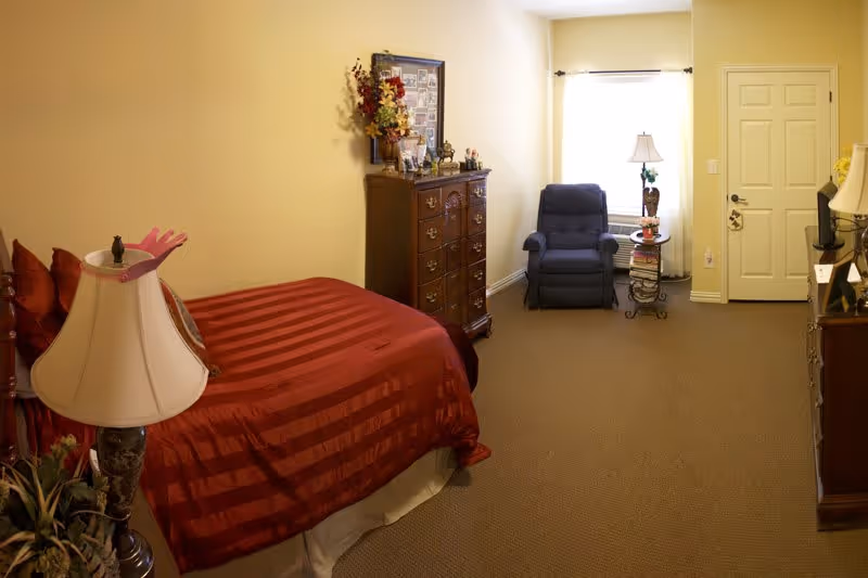 A bedroom with a bed covered in a red striped bedspread, a wooden dresser with decorative items and a framed photo collage on top, a blue recliner chair near a window with white curtains, a side table with a lamp, and a door in the background. The walls are painted light yellow and the floor is carpeted.
